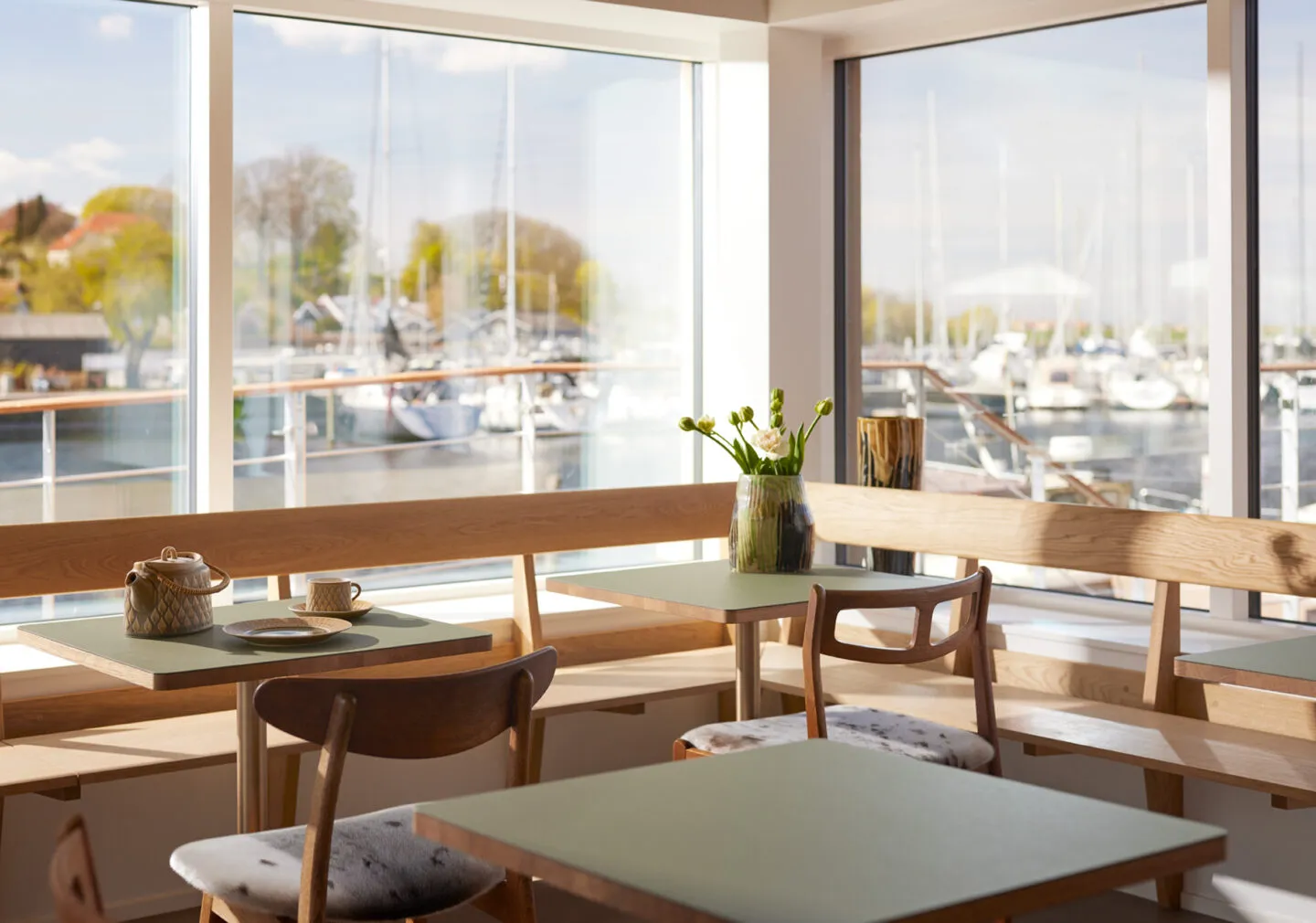 Moderne café indrettet med træborde og stole foran store vinduer med udsigt til bådhavn.