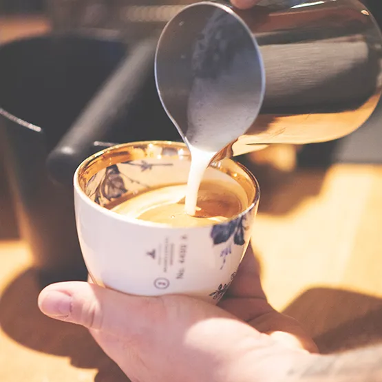 Mælk hældes i en hvid kaffekop med kaffe og latte art.