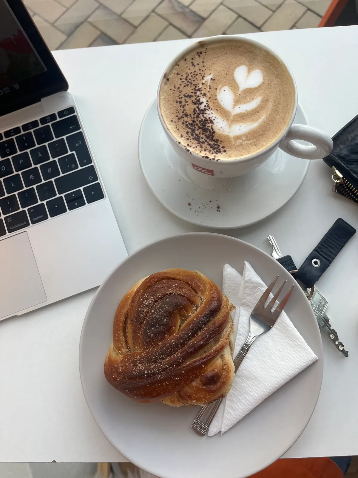 Latte med hjertemønster og friskbagt snegl på hvidt bord med laptop.