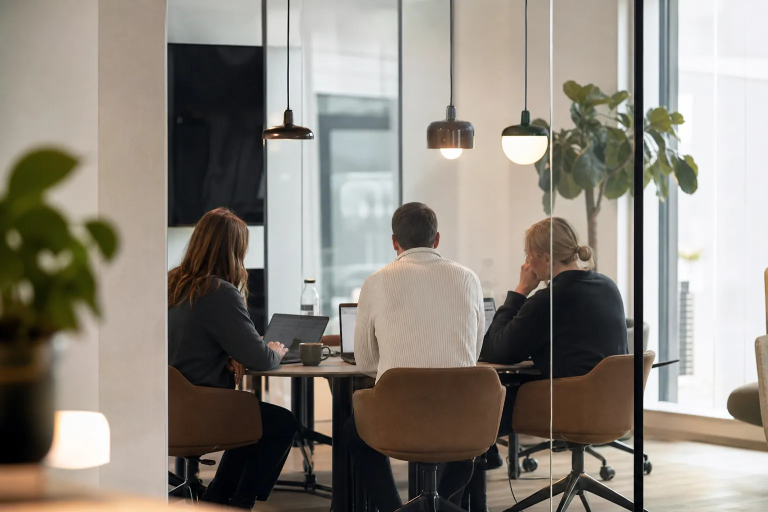 Tre medarbejdere sidder ved et bord i et moderne kontor og arbejder sammen på deres bærbare computere.
