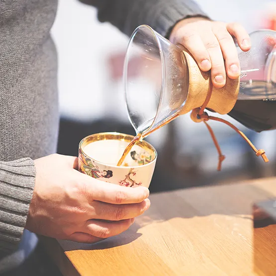 Kaffe bliver hældt fra glaskande ned i et keramisk krus med gulddetaljer.