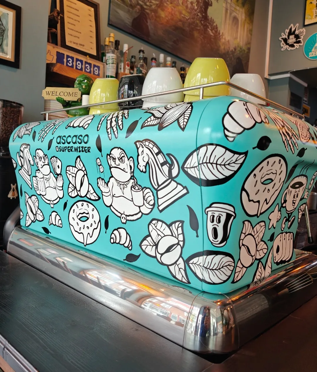 En kunstnerisk dekoreret turkis espressomaskine på en kafé omgivet af farverige kopper.