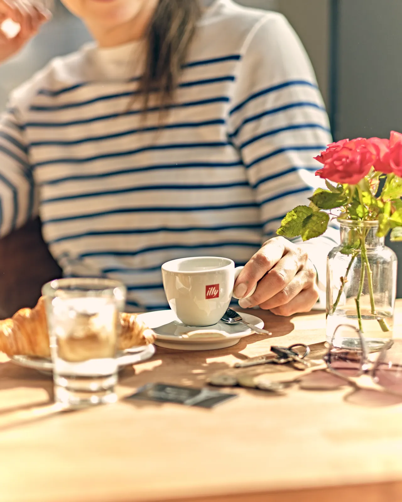 En person i stribet trøje holder en kaffekop ved et bord med blomster og morgenbrød i café-atmosfære.